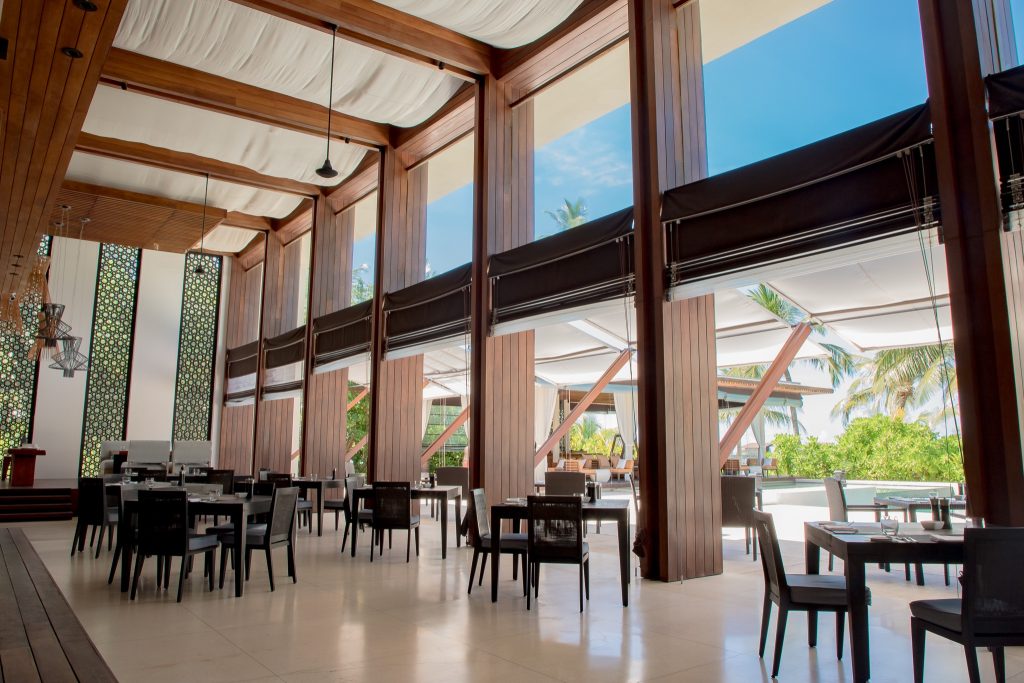Innenansicht des Restaurants The Dining Room im Park Hyatt Maldives Hadahaa mit Holzelementen und Blick auf tropische Palmen