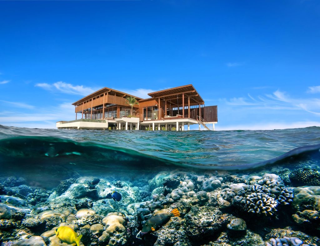 Hotelgebäude auf Holzpfählen über türkisfarbenem Wasser mit Korallenriffen und blauem Himmel auf den
