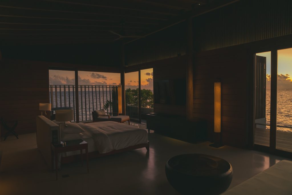 Hotelzimmer mit Holzwänden, großem Bett, Fensterfront und Terrasse mit Meerblick bei Sonnenuntergang