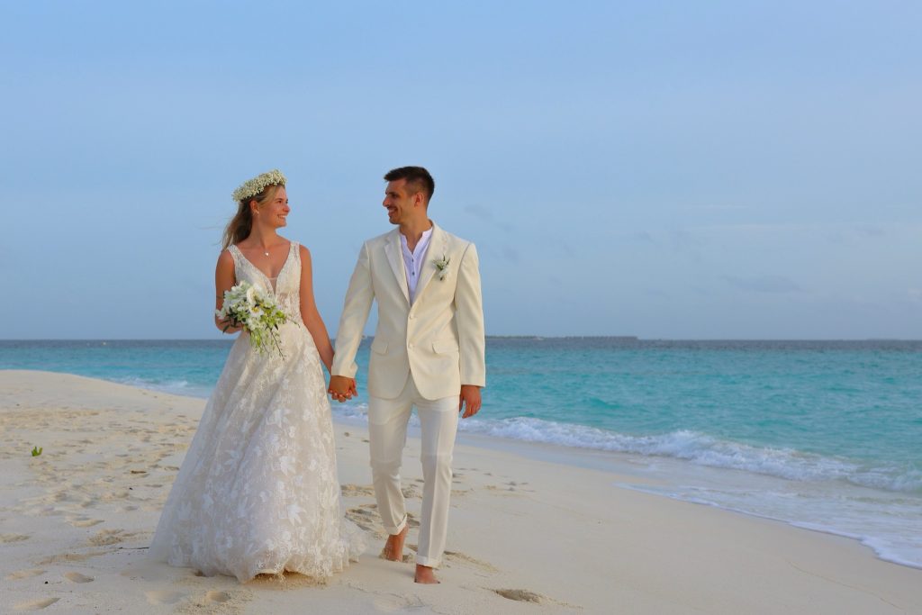 Paar in Hochzeitskleidung hält Händchen am Sandstrand mit türkisfarbenem Meer im Hintergrund