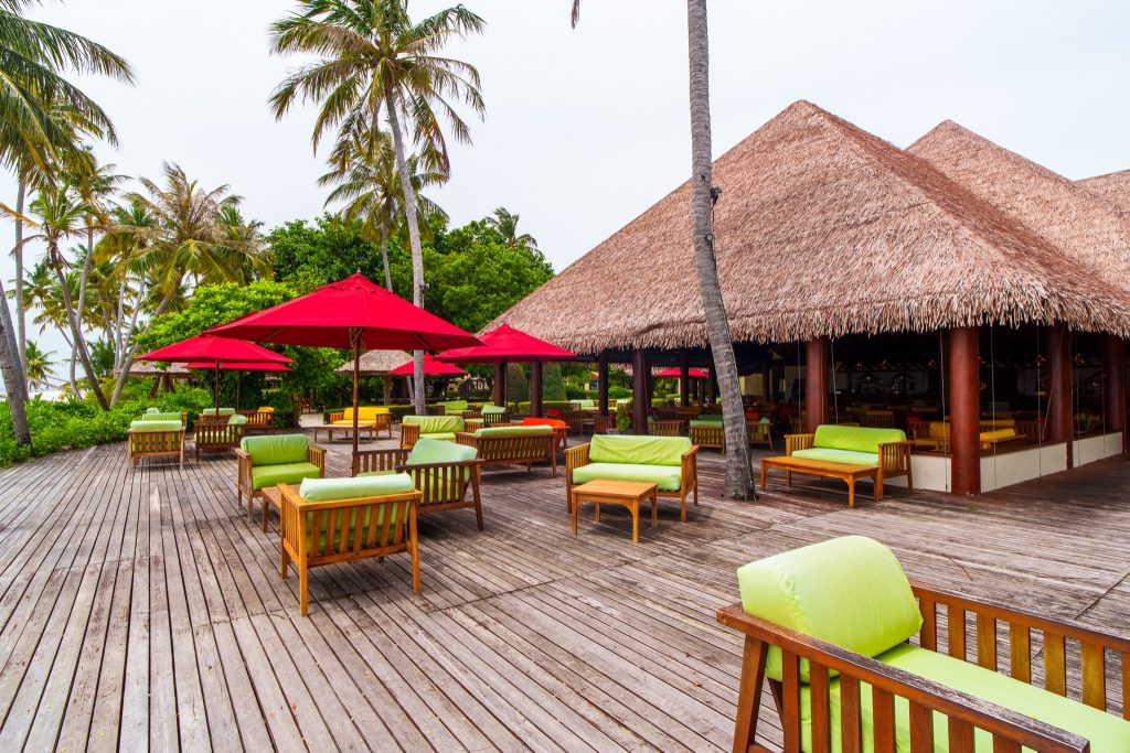 Außenbereich der Haruge Bar im Reethi Faru Resort mit Holzdielen, grünen Sitzpolstern und roten Sonnenschirmen