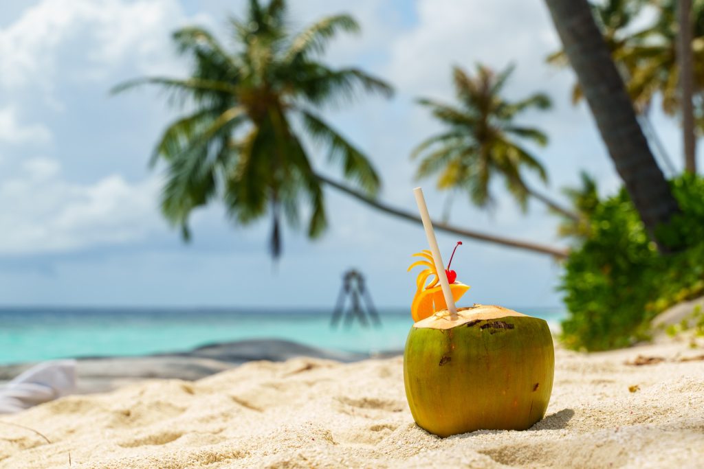 Frische Kokosnuss mit Strohhalm und Fruchtdekoration auf Sandstrand vor türkisfarbenem Meer und Palmen im Reethi Faru Resort.