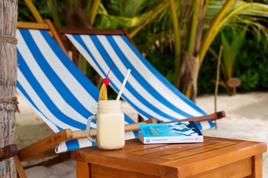 Milchshake mit Kirsche und Bananenstück auf Holztisch vor blau-weiß gestreiften Liegestühlen am Strand im Reethi Faru Resort