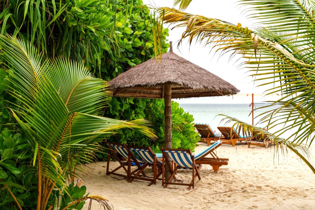 Strandbereich mit blau-weiß gestreiften Liegestühlen unter Strohdach am Meer im Reethi Faru Resort