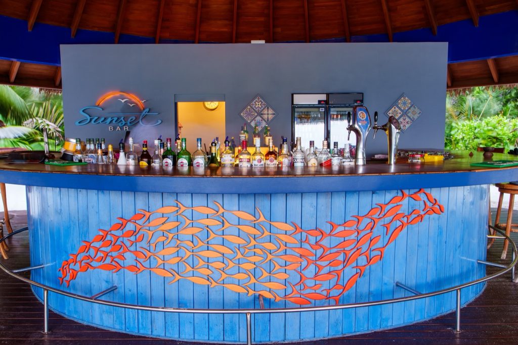 Offene Hotelbar im Reethi Faru Resort mit blauer Theke, orangefarbenen Fischmotiven, Spirituosen und zwei Bierzapfhähnen