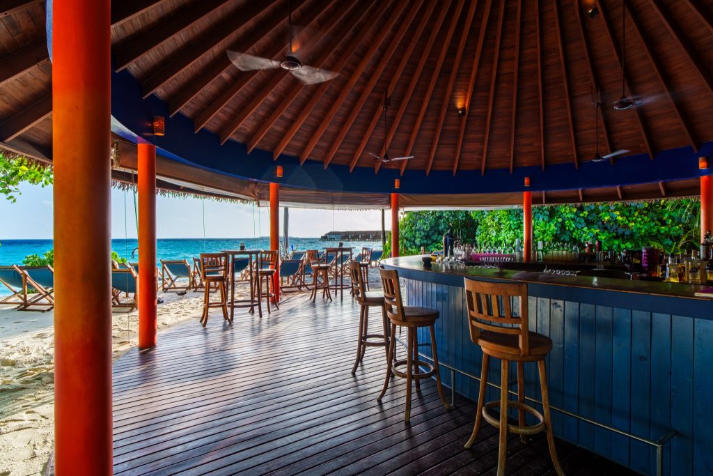Offene Strandbar im Reethi Faru Resort mit blauem Holztresen, orangenen Säulen und Liegestühlen am Meer auf den