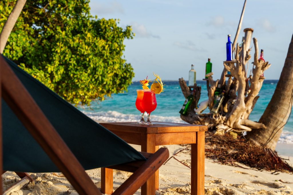 Strand mit hölzernem Tisch, zwei roten Cocktails, Holzliegestuhl, Treibholz mit Glasflaschen und blaugrünem Meer.