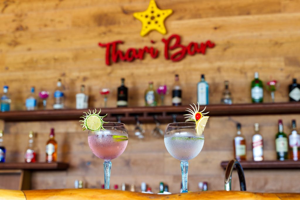 Cocktails auf hölzerner Theke vor Thari Bar mit Holzregalen und Spirituosen im Reethi Faru Resort