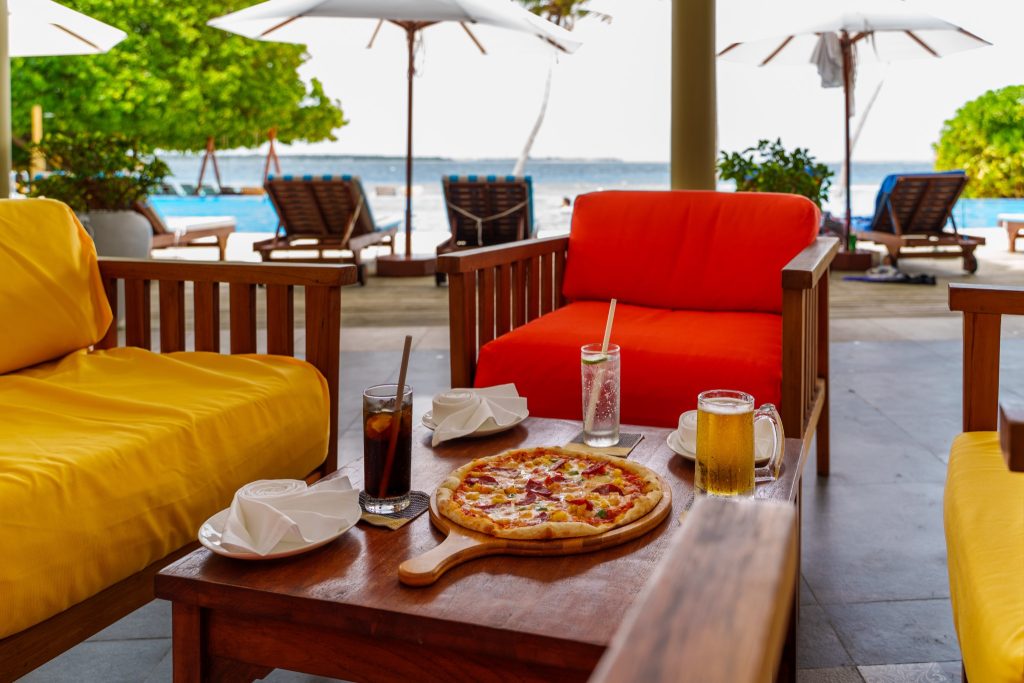 Außenterrasse der Bar Veyo im Reethi Faru Resort mit Holzmöbeln, farbigen Polstern, Pizza und Getränken, Pool und Meer.