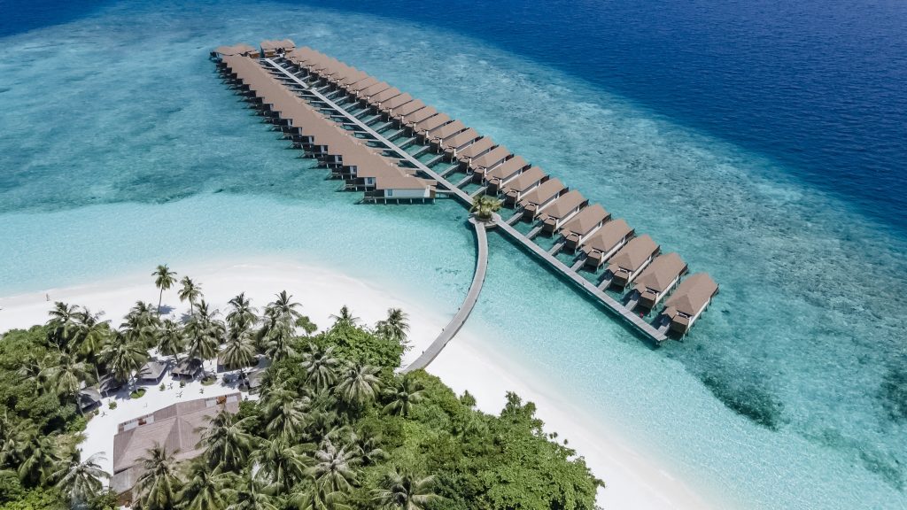Luftaufnahme des Reethi Faru Resorts auf den Malediven mit braunen Überwasser-Bungalows und türkisblauem Wasser
