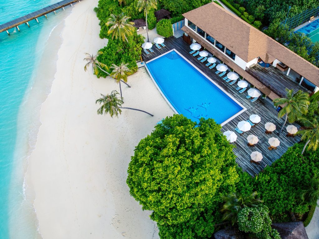 Vogelperspektive auf rechteckigen Pool mit Holzterrasse, Sonnenliegen, Palmen und weißem Sandstrand am Meer