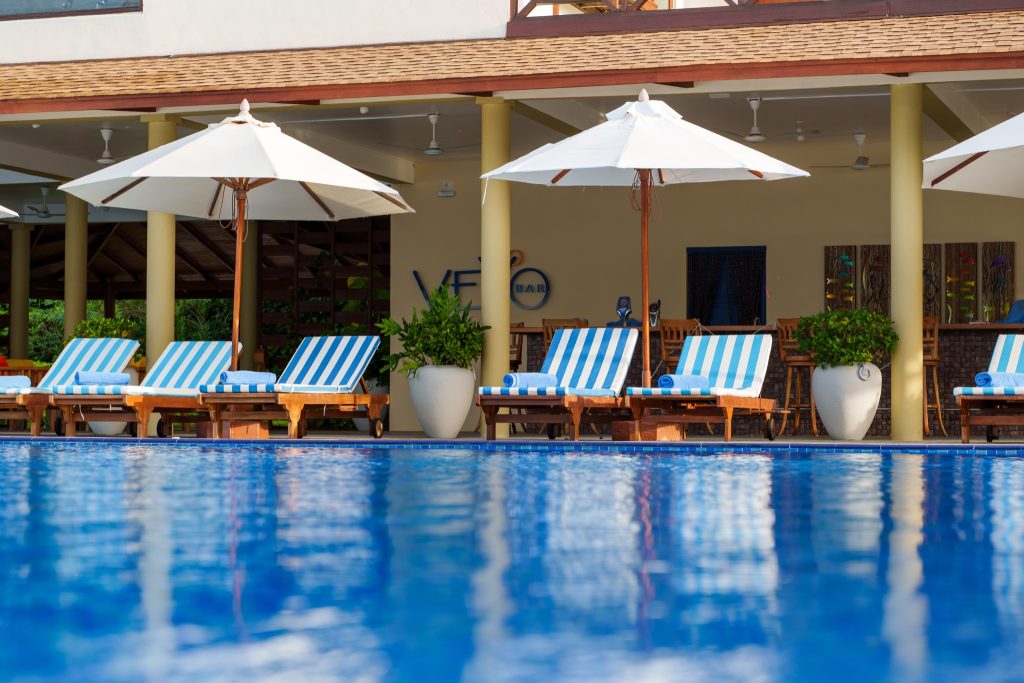 Hotelpool mit blau-weiß gestreiften Liegestühlen, weißen Sonnenschirmen und Holzterrasse im Reethi Faru Resort