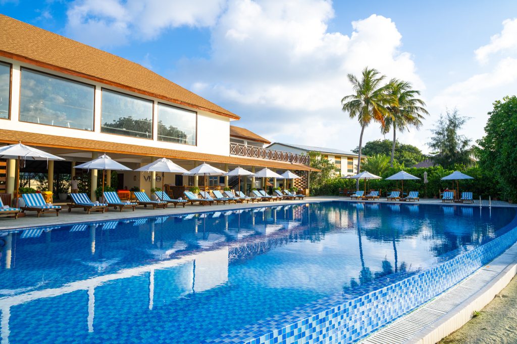 Großer Hotelpool mit blauen und weißen Liegestühlen unter weißen Sonnenschirmen und rotdachigem Gebäude im Hintergrund