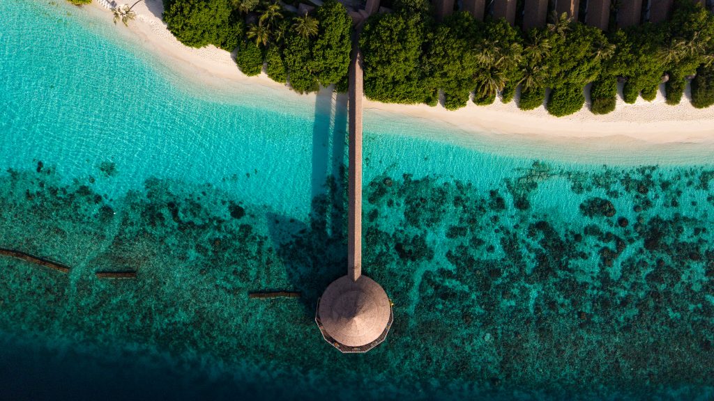 Luftaufnahme vom Restaurant Dhiyavaru im Reethi Faru Resort mit Steg über türkisfarbenem Wasser und rundem Holzpavillon
