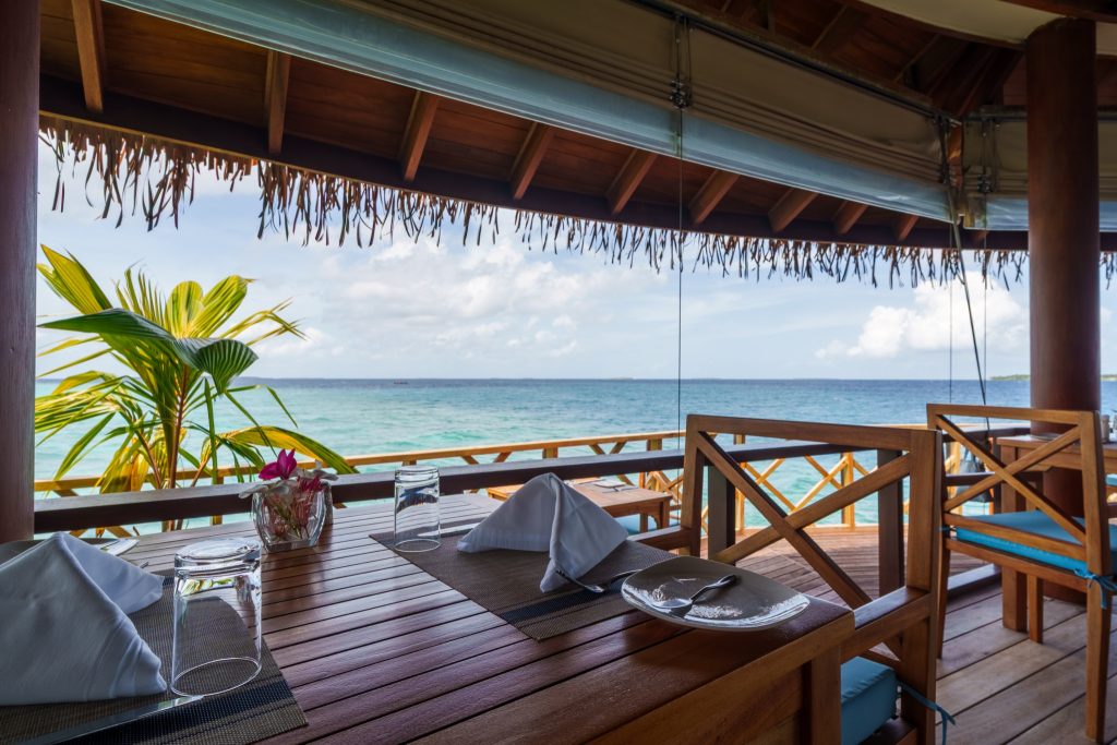 Terrasse des Restaurants Dhiyavaru im Reethi Faru Resort mit Holztisch, weißen Servietten und Blick auf das türkisfarbene.