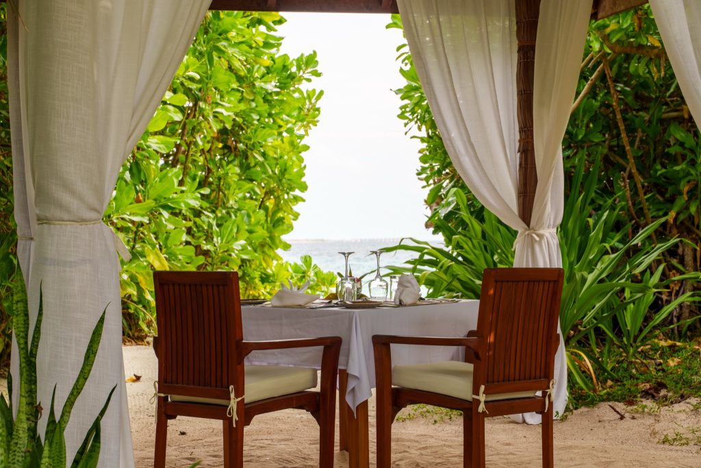 Tisch für zwei unter Pavillon am Strand mit weißen Vorhängen, Holzmöbeln und tropischer Vegetation im Hintergrund