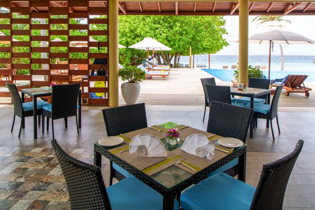 Restaurantbereich Raalhu Cafe mit geflochtenen Möbeln, blauen Kissen, Glas-Tischen und Blick auf Pool und Meer im Reethi.