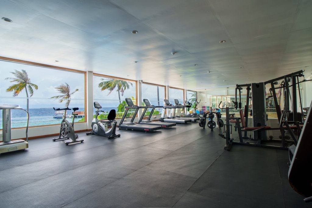 moderner Fitnessraum im Reethi Faru Resort mit Laufbändern, Fahrradergometern, Kraftstationen und Meerblick durch raumhohe.