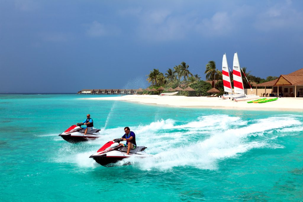 Zwei Personen auf Jetskis fahren über türkisblaues Wasser vor weißem Sandstrand mit Palmen
