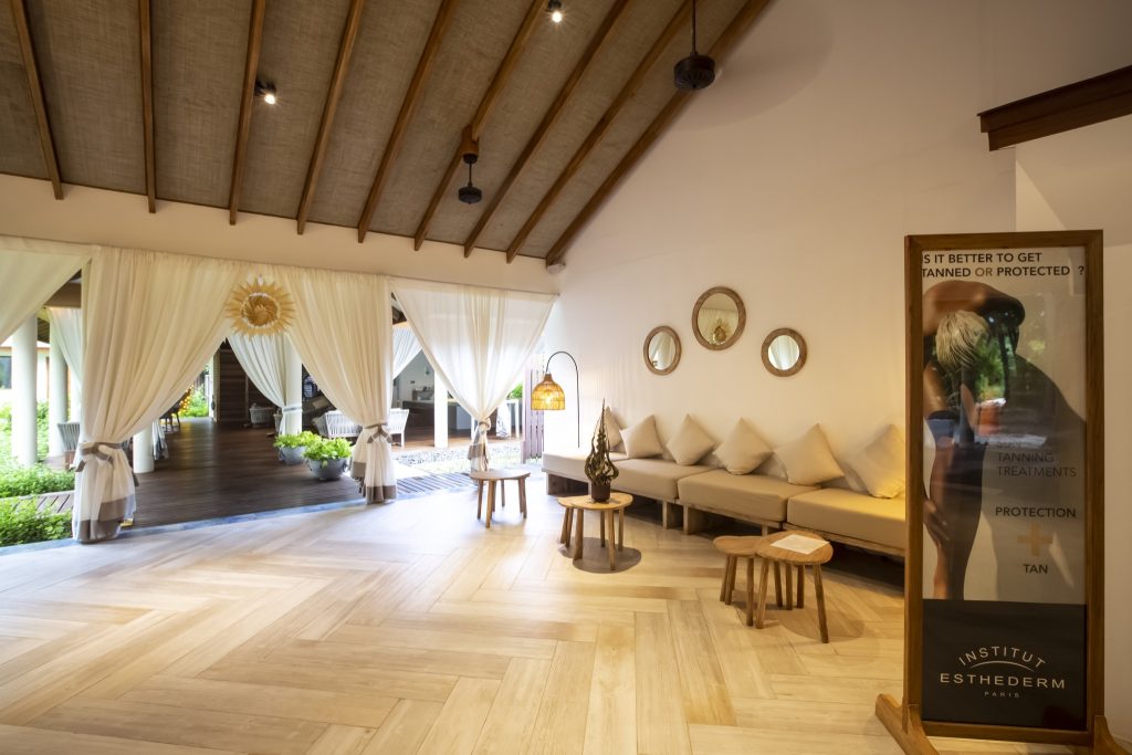 Innenbereich der Wellness Coconut Spa Lounge mit Sitzmöbeln, Holztischen, Fischgrätboden und Blick auf Außenbereich