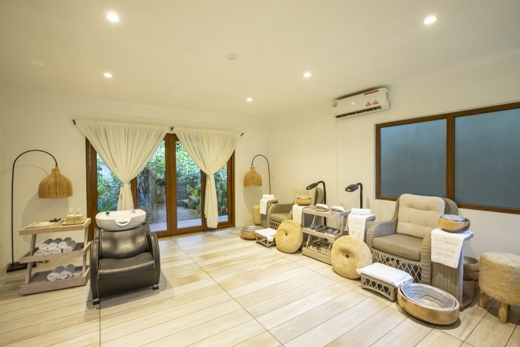 Wellnessbereich im Reethi Faru Resort mit Friseurstuhl, Waschbecken, Rattanmöbeln und großen Fenstern mit weißen Vorhängen