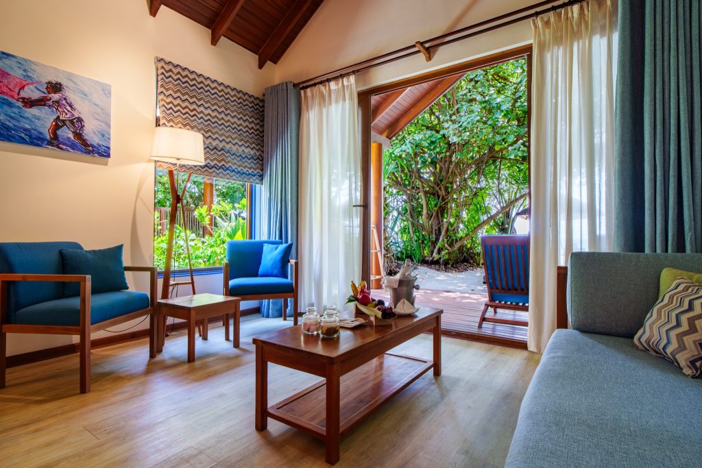 Modernes Deluxe Beach Villa Zimmer im Reethi Faru Resort mit Terrasse, blauen Sesseln, Holztisch und frischem Obst