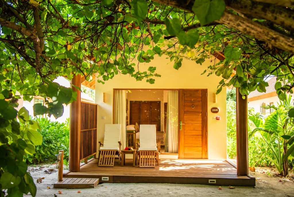 Vorderansicht der Garden Villa im Reethi Faru Resort mit Holzterrasse, zwei Sonnenliegen und üppigem Grün