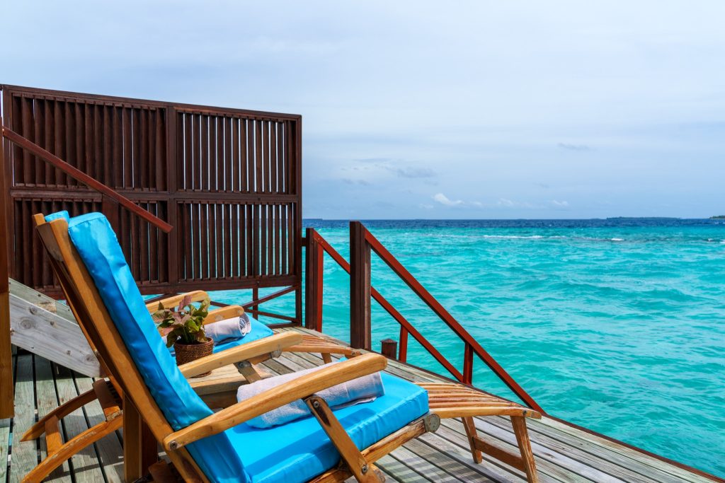 Terrasse der Water Villa im Reethi Faru Resort mit zwei Holzliegestühlen, blauem Kissen, kleinem Tisch und Meerblick