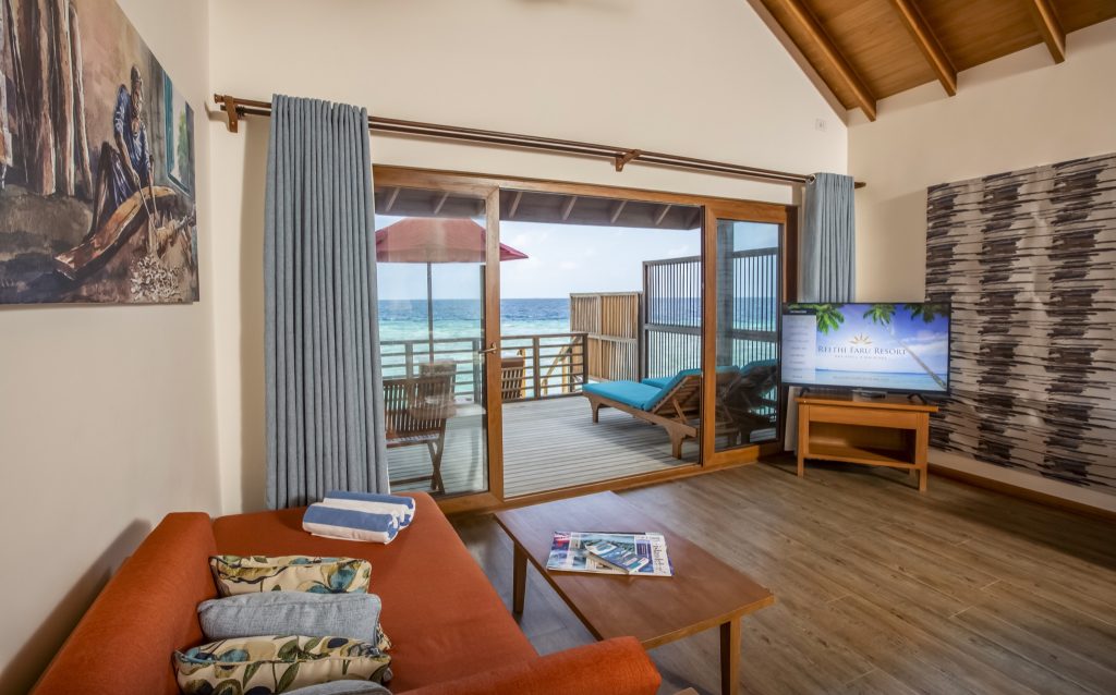 Zimmer Water Villa im Reethi Faru Resort mit orangefarbenem Sofa, Holzboden und Balkon mit Meerblick
