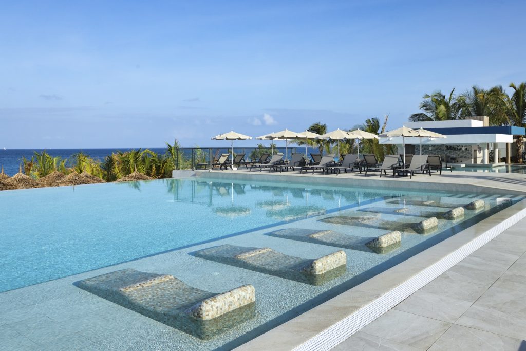 Infinity-Pool mit gekachelten Liegen im Wasser, Terrasse mit Sonnenliegen und Meer im Hintergrund im Hotel Riu Palace Swahili