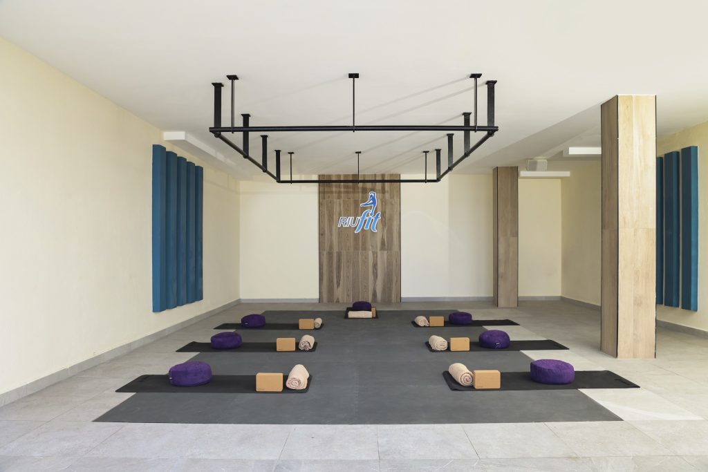 Moderner Fitnessraum Hotel Riu Palace Swahili mit Yogamatten, Kissen, Yogablöcken und Holzpaneelen mit RIU fit Logo