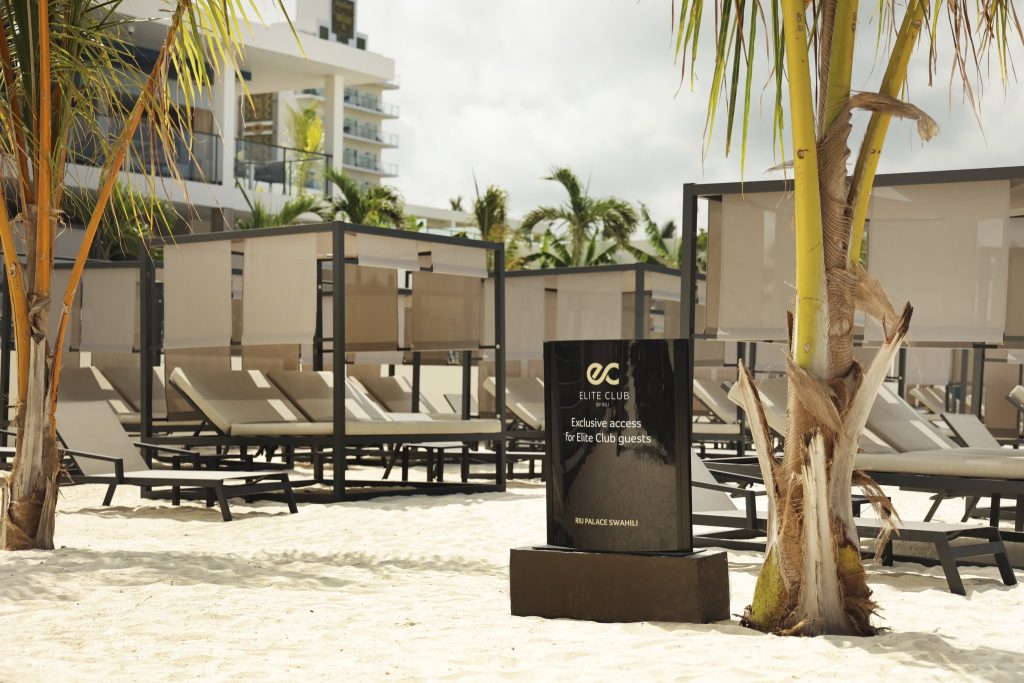 Strandbereich des Hotel Riu Palace Swahili mit Elite Club Schild, Palmen, Liegen und rechteckigen Sonnenschirmen