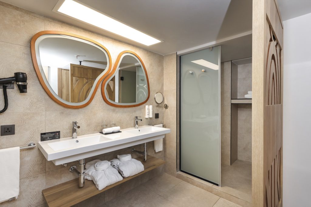 Modernes Badezimmer mit zwei asymmetrischen Holzspiegeln, Doppelwaschbecken und Holzregal im Hotel Riu Palace Swahili