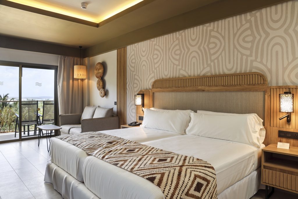 Modernes Hotelzimmer Junior Suite Twin mit Doppelbett, Holzelementen, beige Tapete und Sitzgruppe am Fenster im Hotel Riu.