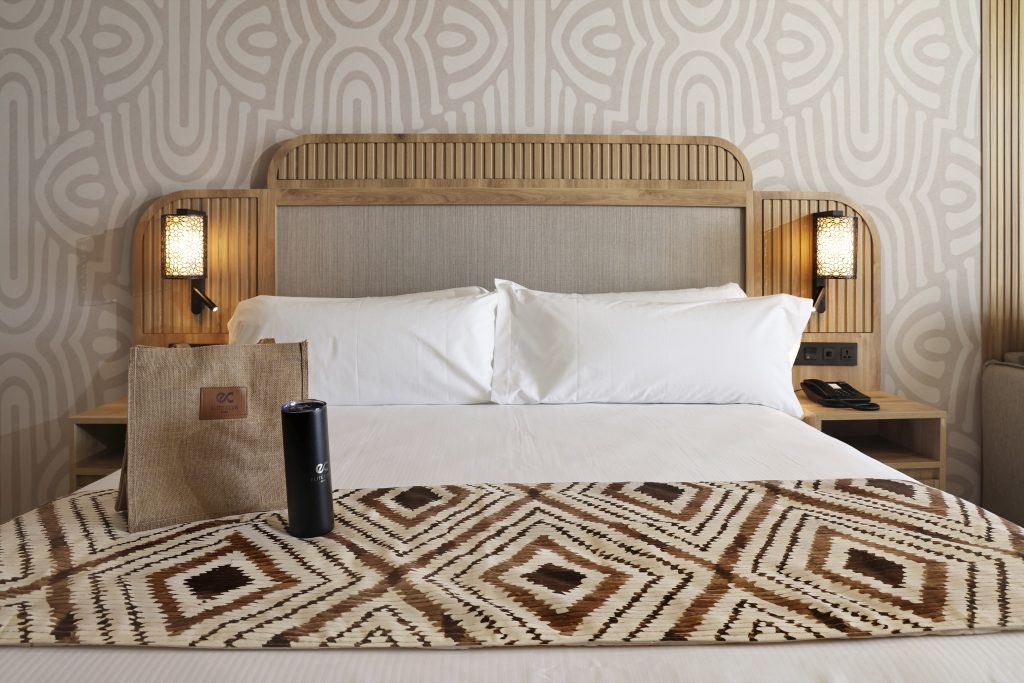 Hotelzimmer mit großem Bett, weißer Bettwäsche, Holz-Kopfteil, zwei Metall-Wandleuchten und geometrischer Tapete