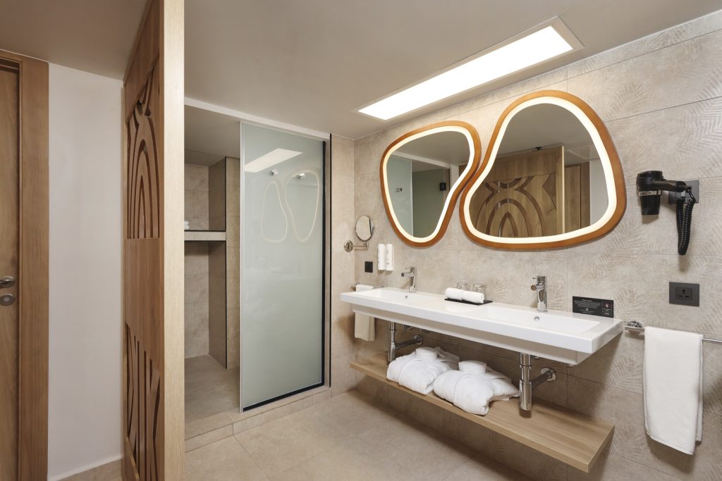 Modernes Badezimmer mit weißen Keramikwaschtisch, asymmetrischen Spiegeln und Holzelementen im Hotel Riu Palace Swahili