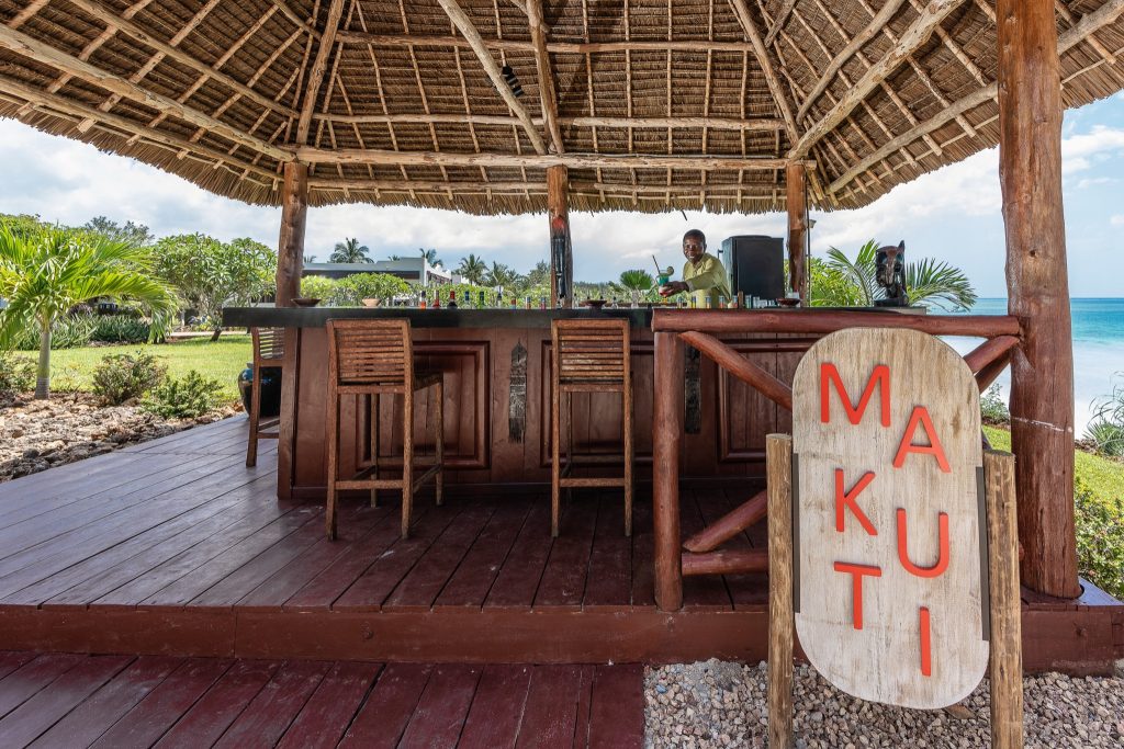 Offene Strandbar Makuti mit Strohdach und Holzhockern, tropische Pflanzen und Meer im Hintergrund