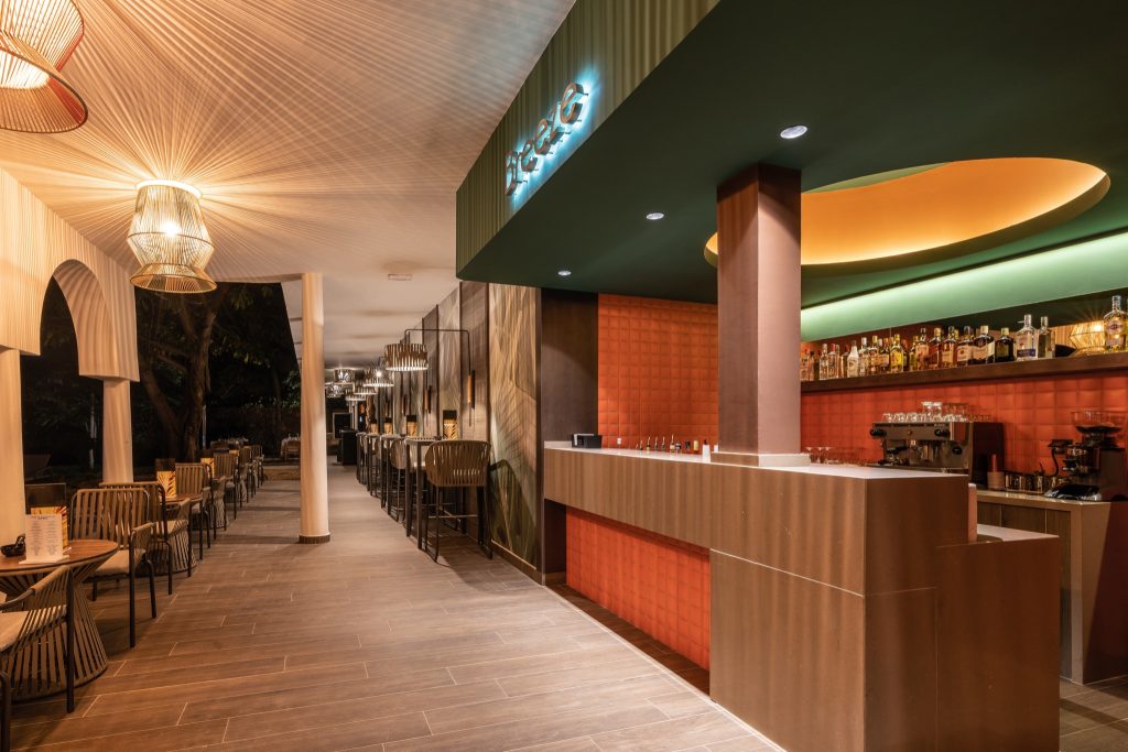 Hotelbar mit rostfarbenem Tresen, Holztischen, geflochtenen Stühlen und warmen Lampen im Riu Palace Zanzibar