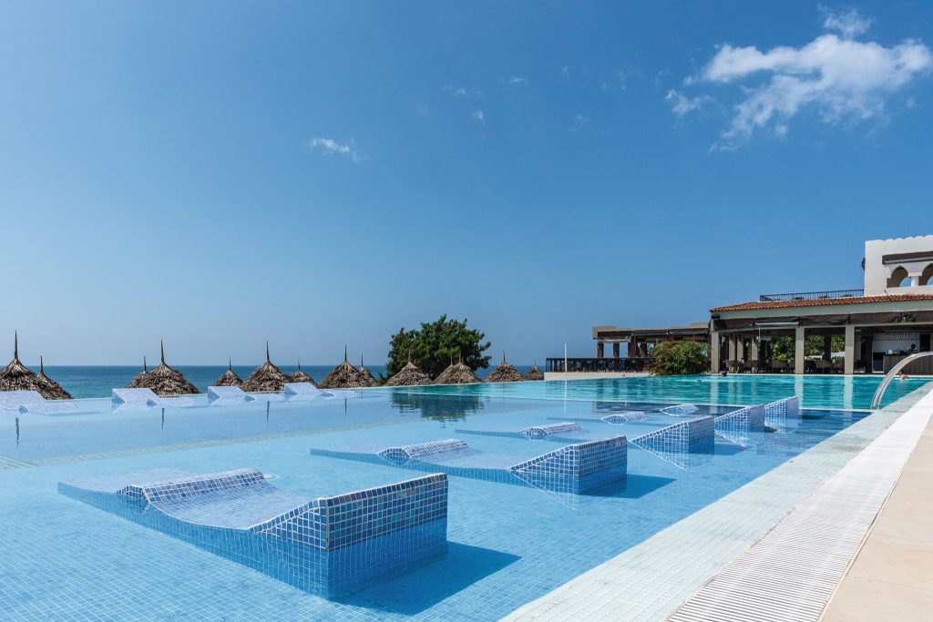 Großer Infinity-Pool mit blauen Mosaikliegen im Wasser und Stroh-Sonnenschirmen am Meer in Sansibar