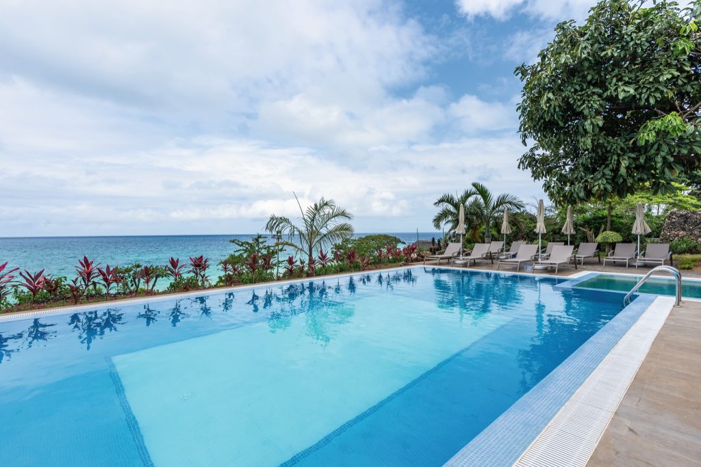 Pool mit klarem blauem Wasser, Holzterrasse, Liegestühlen und Palmen im Hintergrund am Riu Palace Zanzibar
