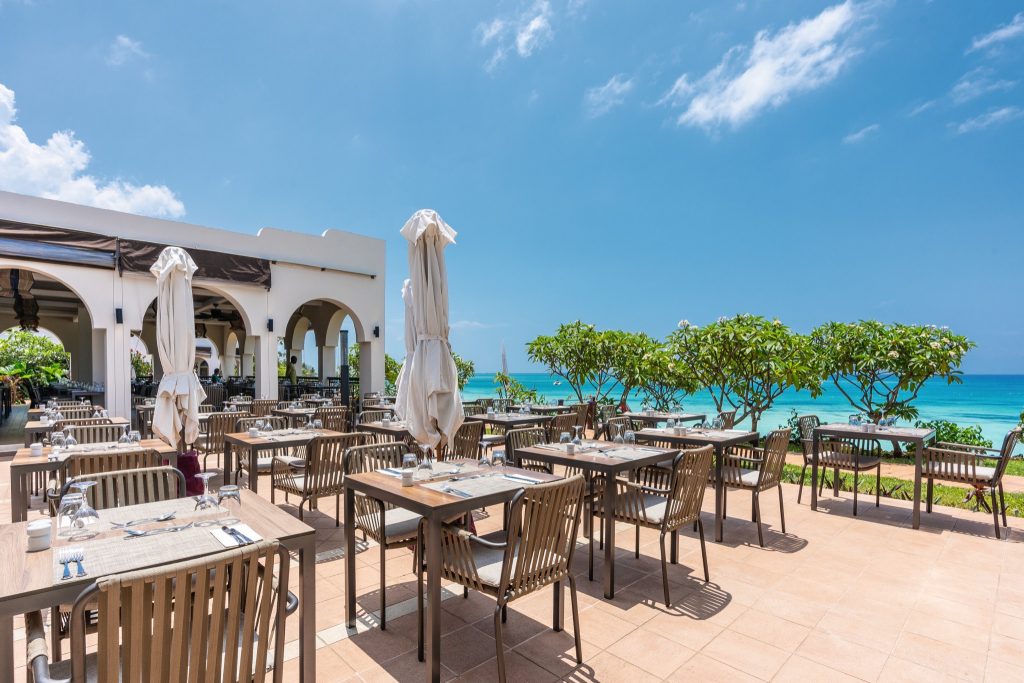 Außenterrasse des Riu Palace Zanzibar Restaurants mit Holzstühlen, Tischen und Sonnenschirmen am Meer