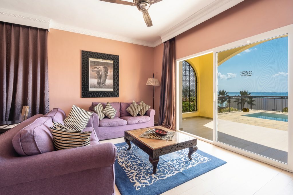 Wohnzimmer der Beach Villa im Riu Palace Zanzibar mit lilafarbenen Sofas, Holztisch, Glas-Schiebetür und Meerblick