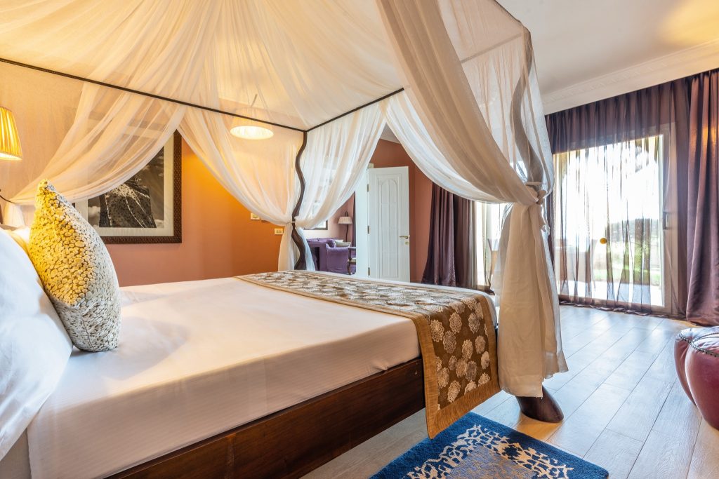 Hotelzimmer mit Himmelbett, weißen Stoffvorhängen, terrakottafarbenen Wänden und Holzboden im Riu Palace Zanzibar