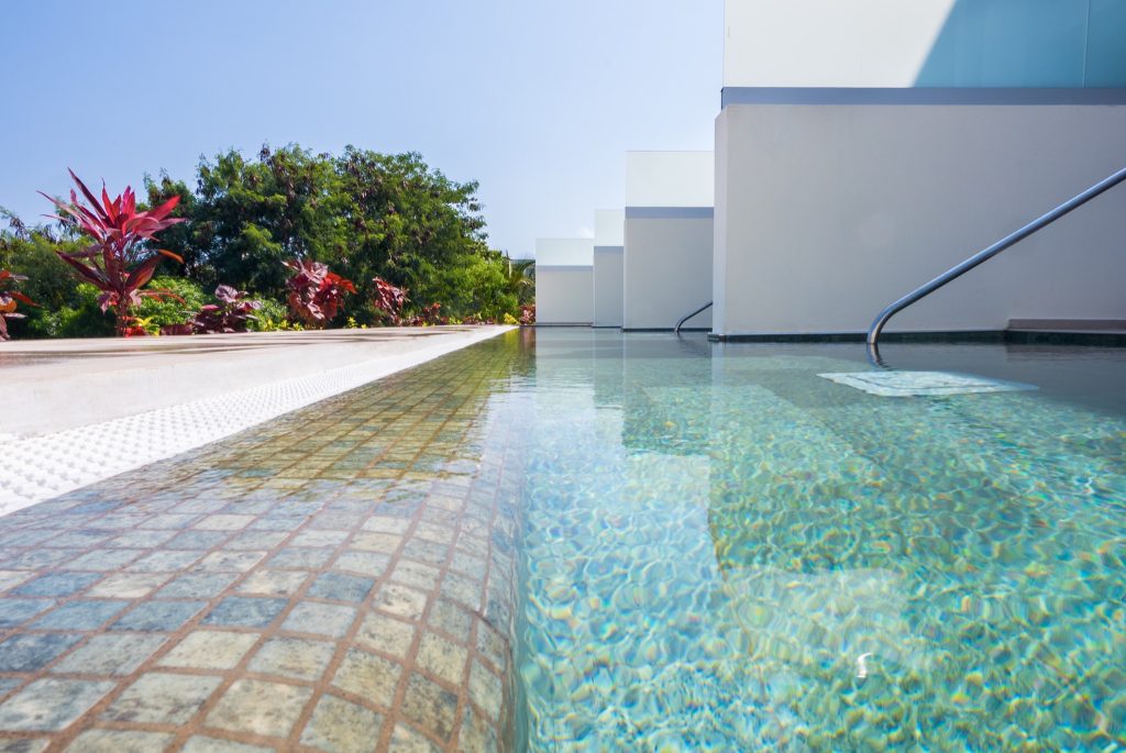 Moderner Pool mit blau-grün gefliesten Boden, weißen Wänden, Metallgeländern und tropischen Pflanzen im Außenbereich