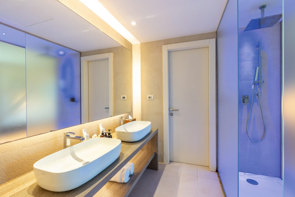 Modernes Badezimmer mit zwei weißen Waschbecken auf Holzablage, großem Wandspiegel und gläserner Duschkabine im Riu Palace.