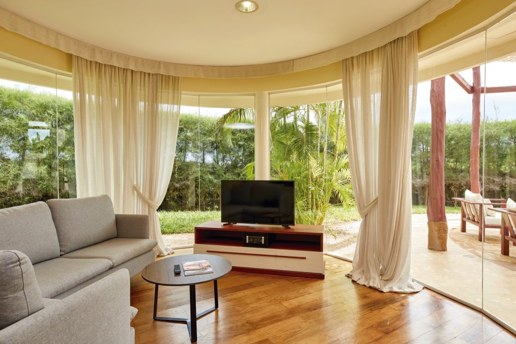 Hotelzimmer im Riu Palace Zanzibar mit großer Glasfront, grauem Sofa, Holztisch und Blick auf grüne Gartenlandschaft