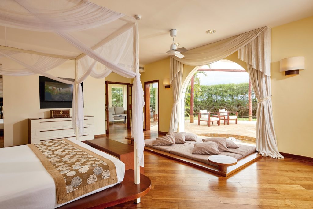 Geräumiges Zimmer im Riu Palace Zanzibar mit Himmelbett, weißer Kommode, Fernseher und Terrasse mit Gartenblick