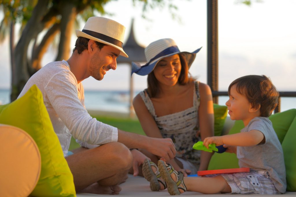 Zwei Erwachsene und ein Kind auf heller Sitzgruppe mit grünen Kissen, Palmen und Meer im Hintergrund