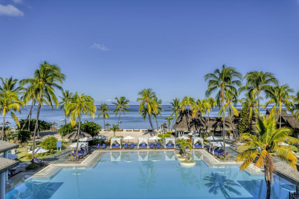 Großer Swimmingpool mit Palmen und Strohdach-Pavillons, Strand und Meer im Hintergrund bei Tageslicht