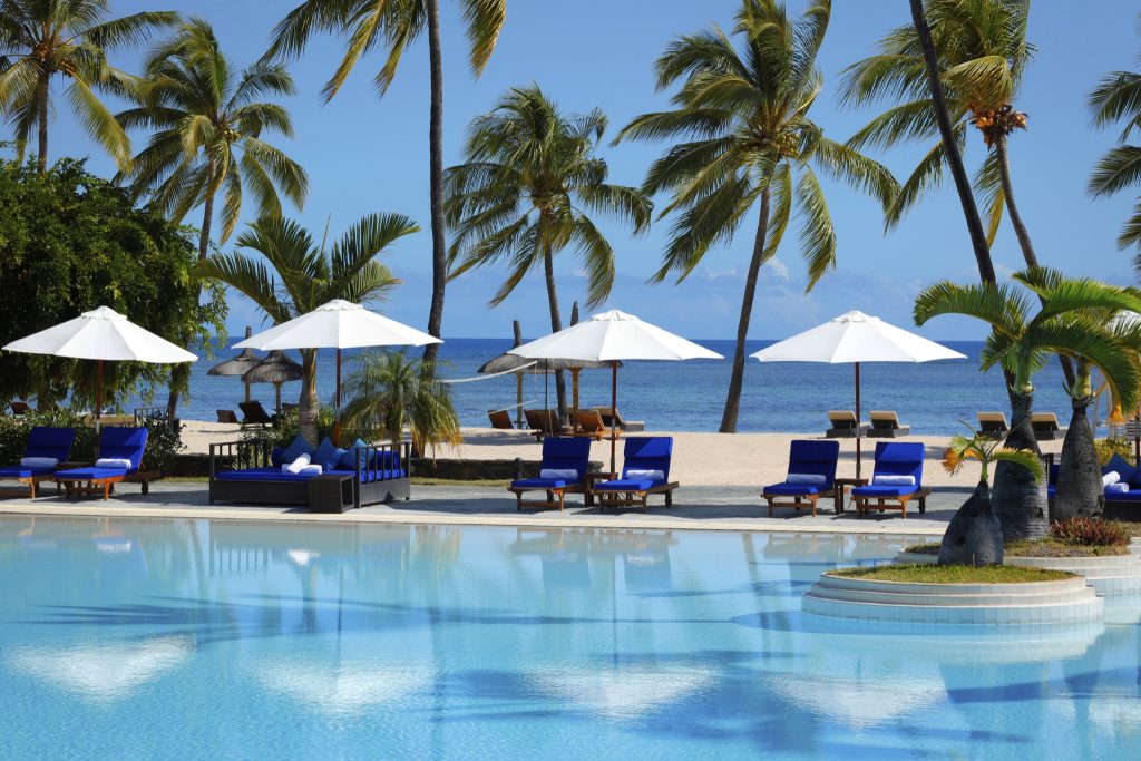 Großer Pool mit blauen Liegestühlen und weißen Sonnenschirmen, Palmen und Meer im Hintergrund